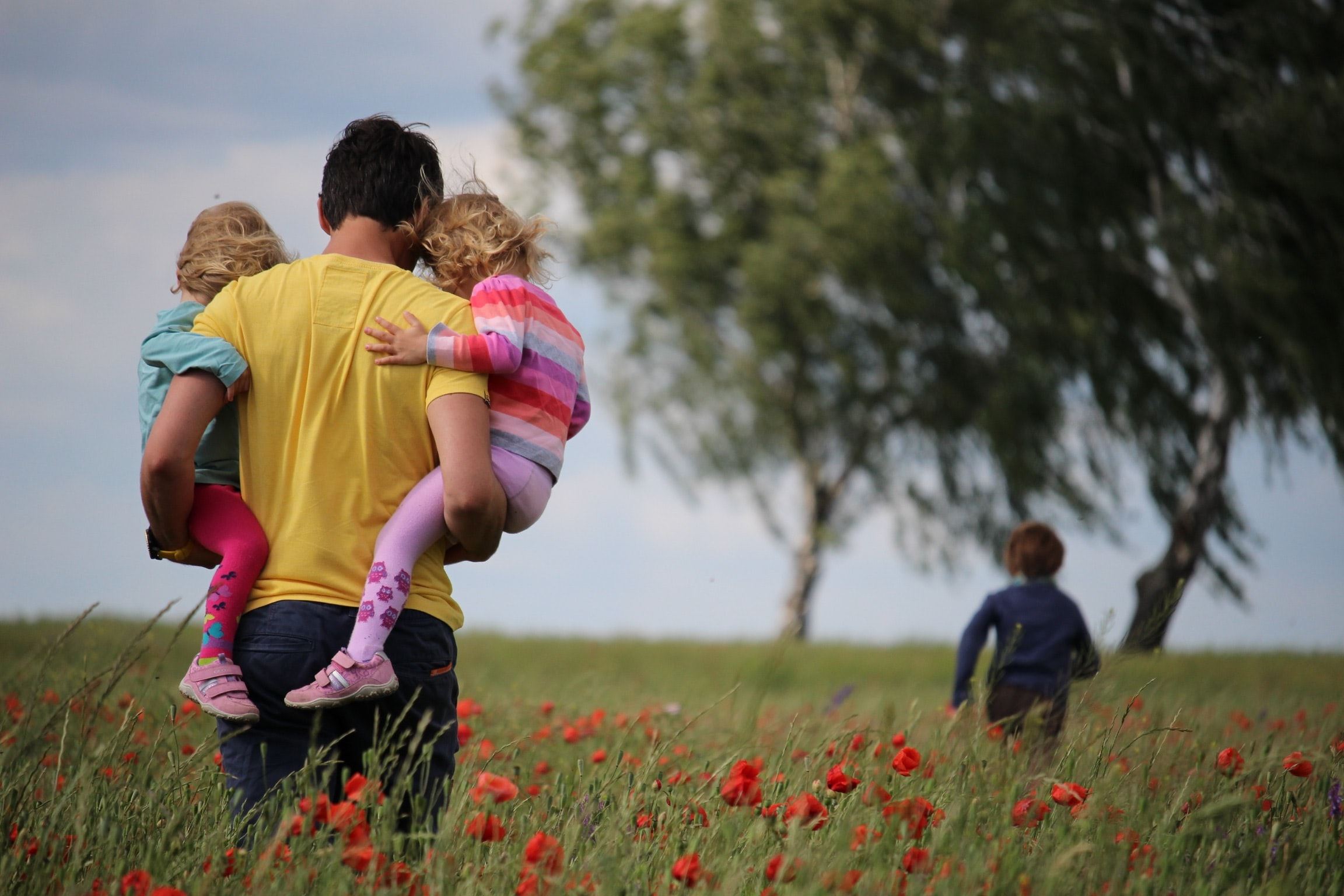 father walking holding two kids while another kid runs ahead